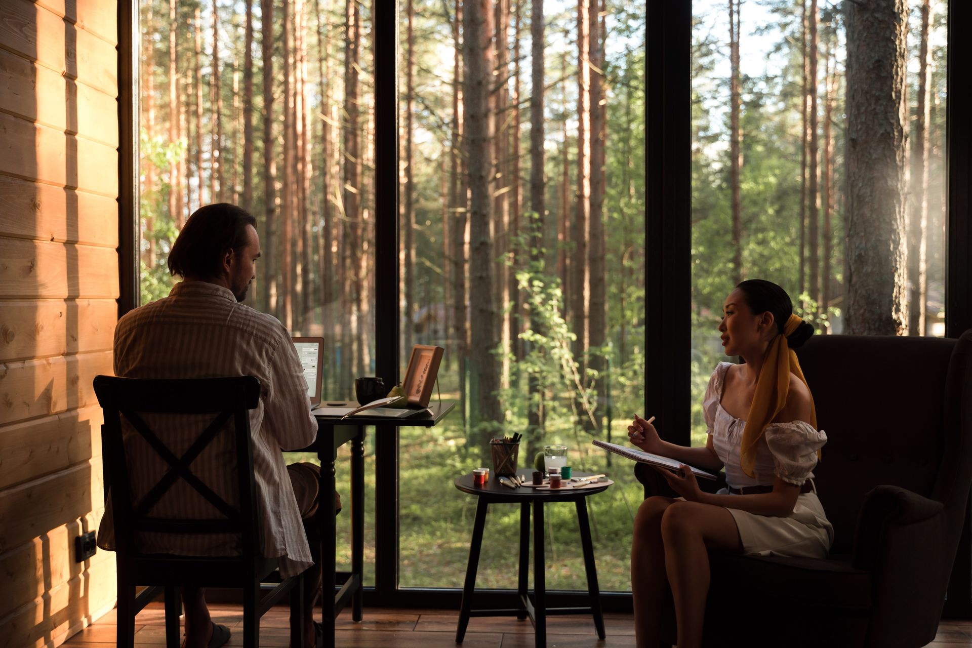 A couple working remotely in a cozy cabin with a forest view through large windows.