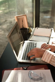 A person typing on a laptop at a sunlit desk with documents and a notebook.