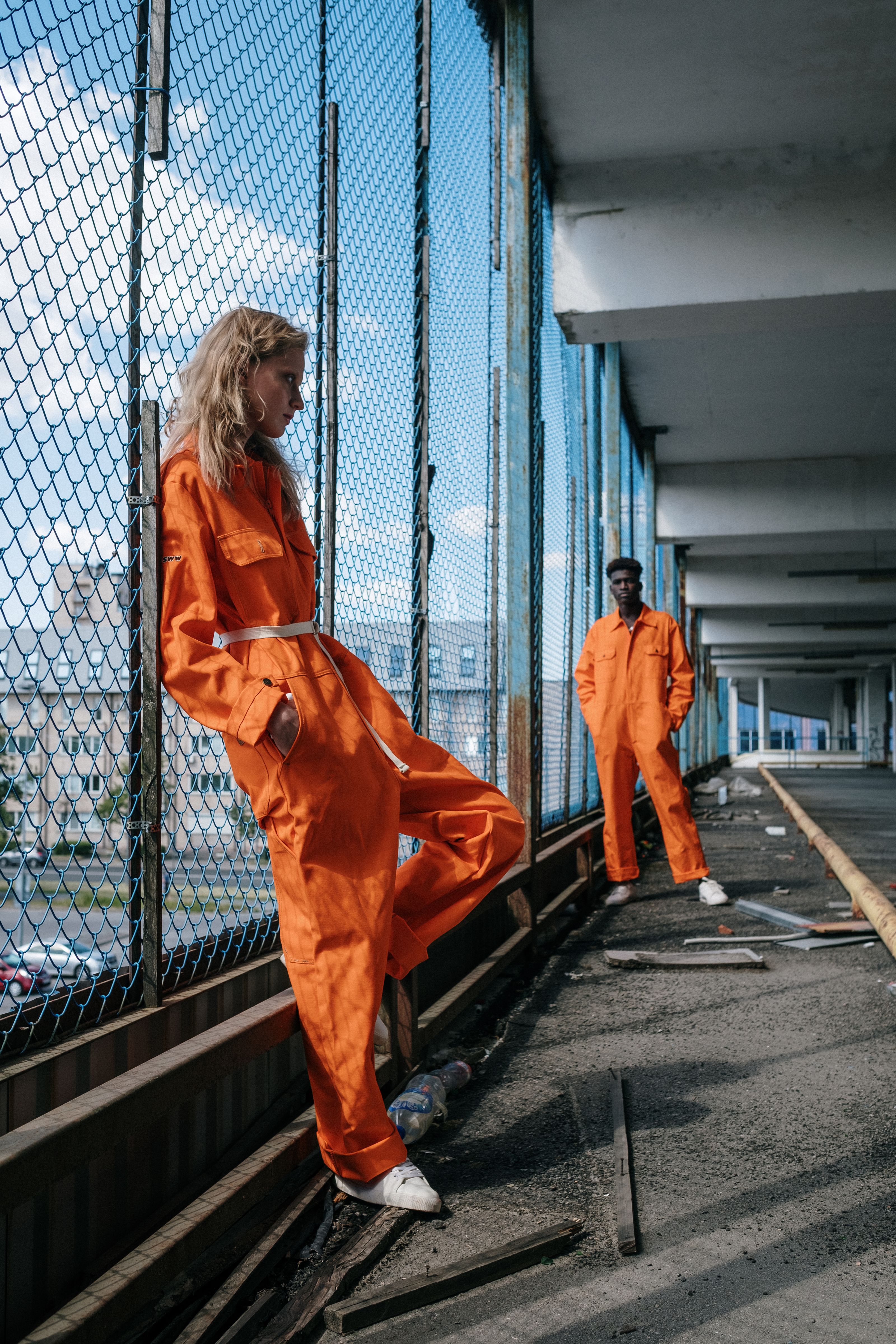 Dois modelos em macacões laranja posam ao lado de uma cerca de cadeado em um ambiente urbano.