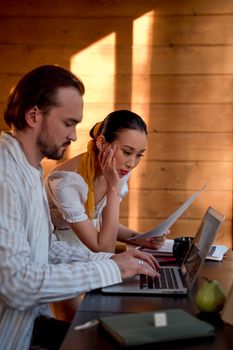 Two adults collaborating on a project using a laptop and papers in a sunlit wooden room.