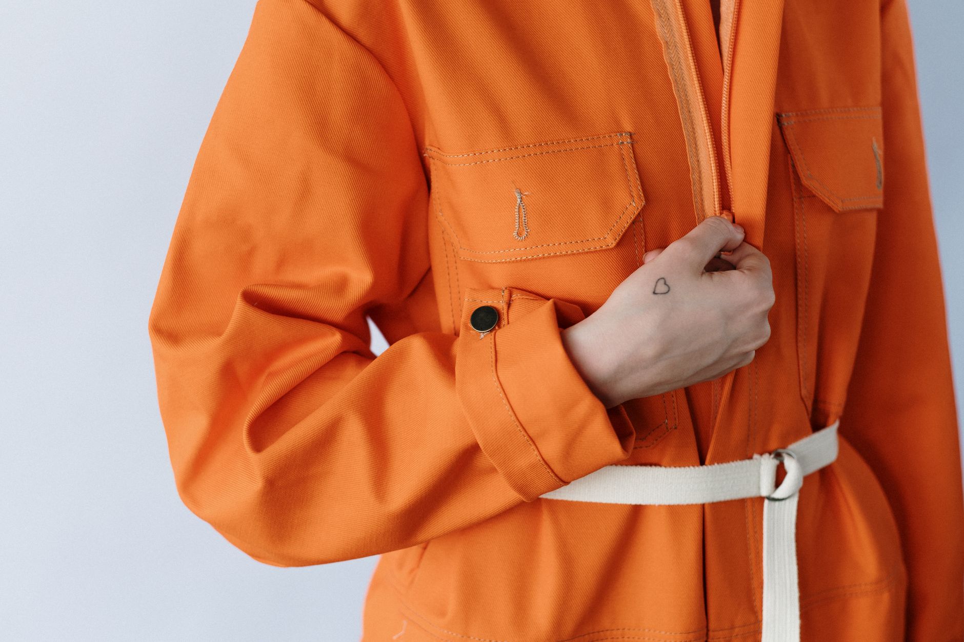 Close-up photo of a person in a stylish orange jumpsuit, highlighting a small heart tattoo on their hand.