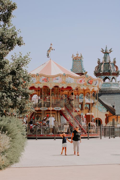 Free A mother and daughter enjoying a walk near a vibrant carousel on a sunny day at an amusement park. Stock Photo