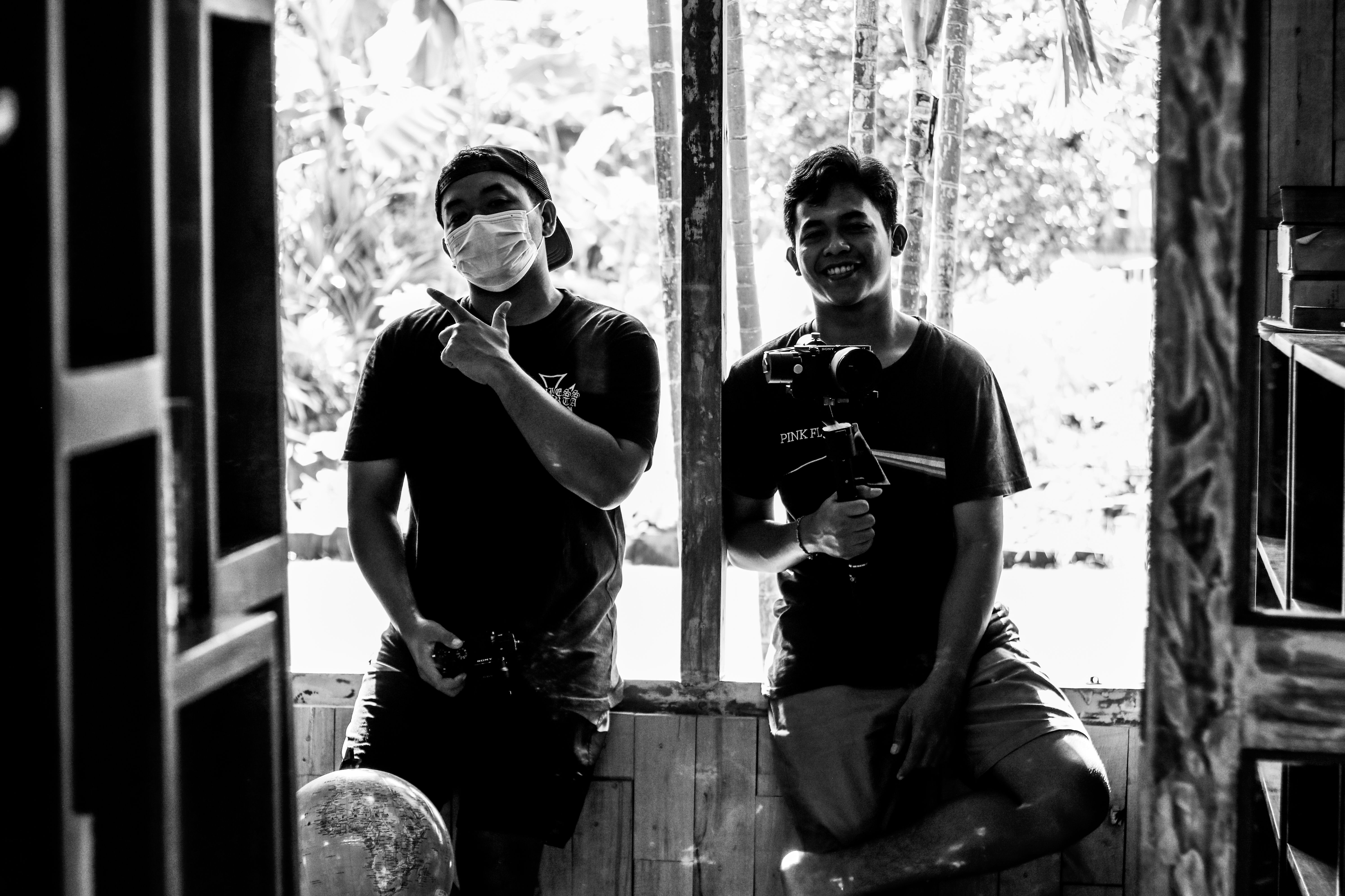 Two men indoors, one smiling while holding a camera, captured in a candid black and white photograph.