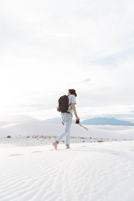 A man walking barefoot with a backpack across the tranquil White Sands dunes under a bright sky.