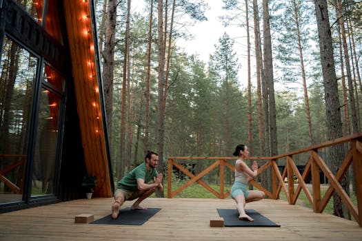 A couple practicing yoga on a wooden deck surrounded by a serene forest setting.