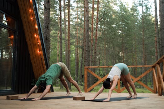 A couple performs yoga on a cabin deck nestled in a serene forest setting.