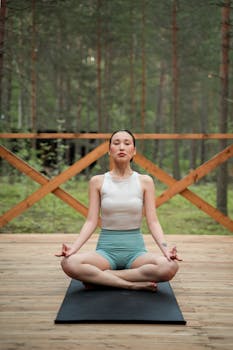A woman meditates in a serene forest setting, practicing yoga on a wooden deck.