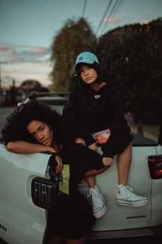 Two young women in streetwear pose on a car during sunset, showcasing trendy fashion.