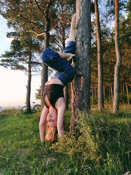 Woman in casual attire doing a handstand against a tree in a sunlit forest setting.