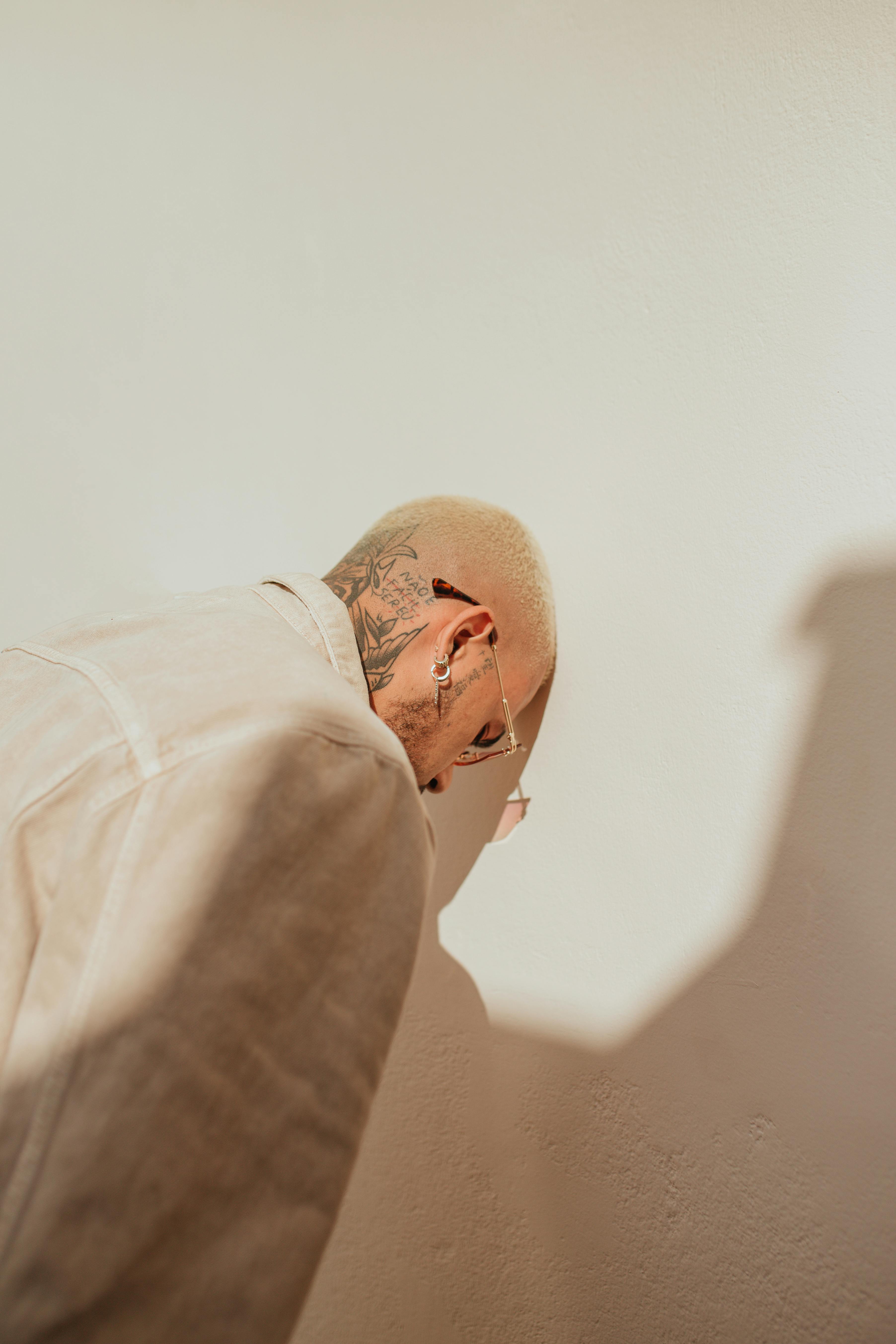 A tattooed man leans against a white wall in a studio, evoking a sense of introspection.