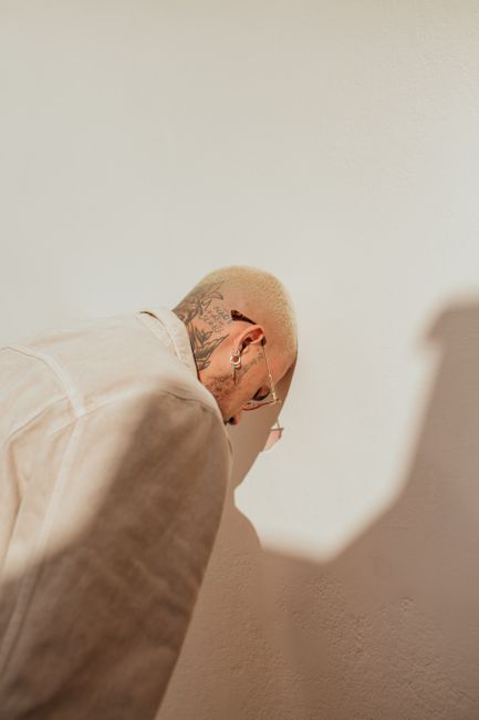 A tattooed man leans against a white wall in a studio, evoking a sense of introspection.