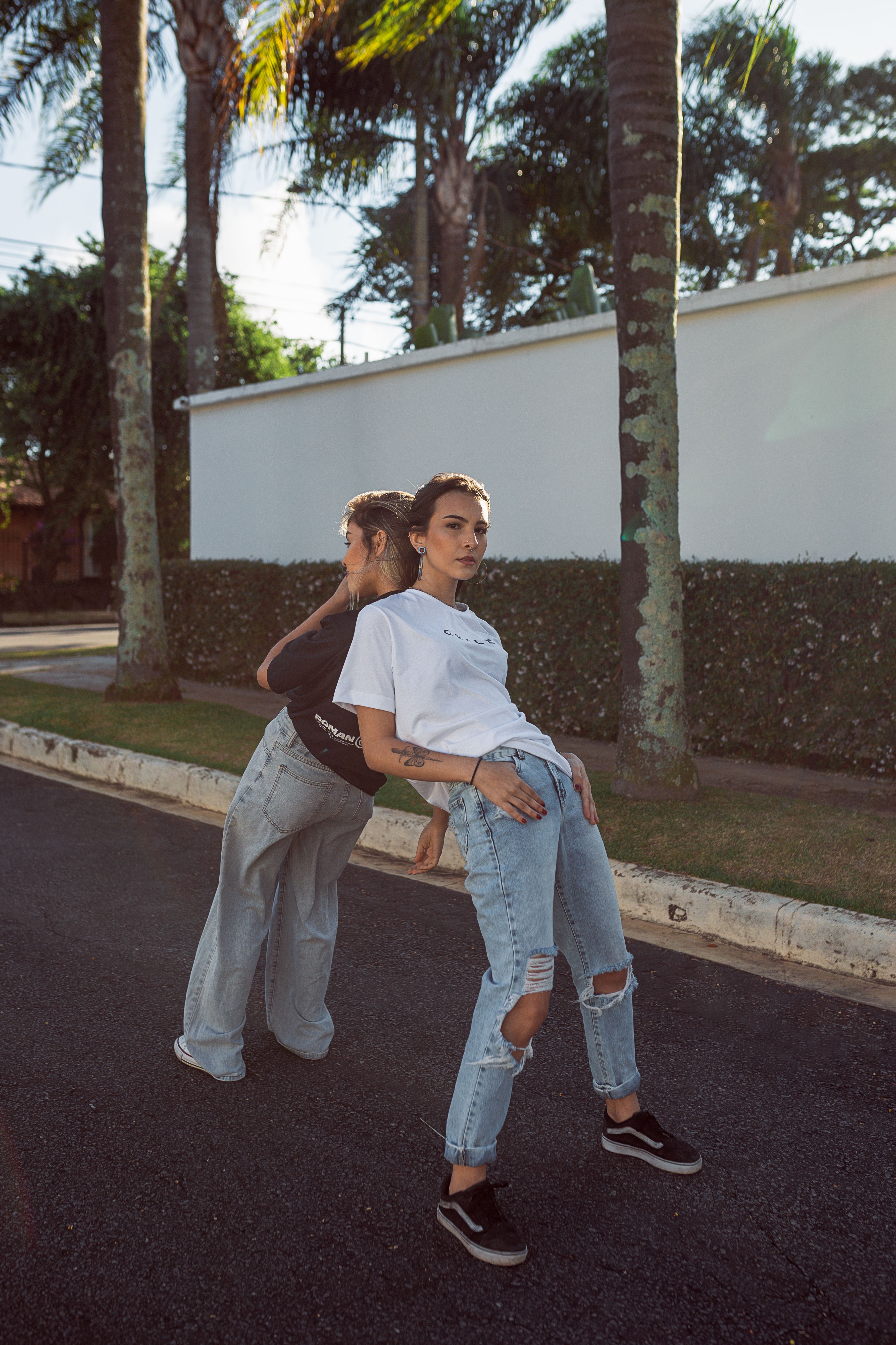 Two stylish women leaning back-to-back on a sunny street, amidst palm trees.