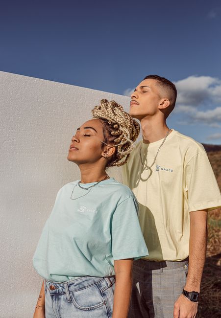 A stylish couple with closed eyes standing outdoors against a white wall in pastel t-shirts.