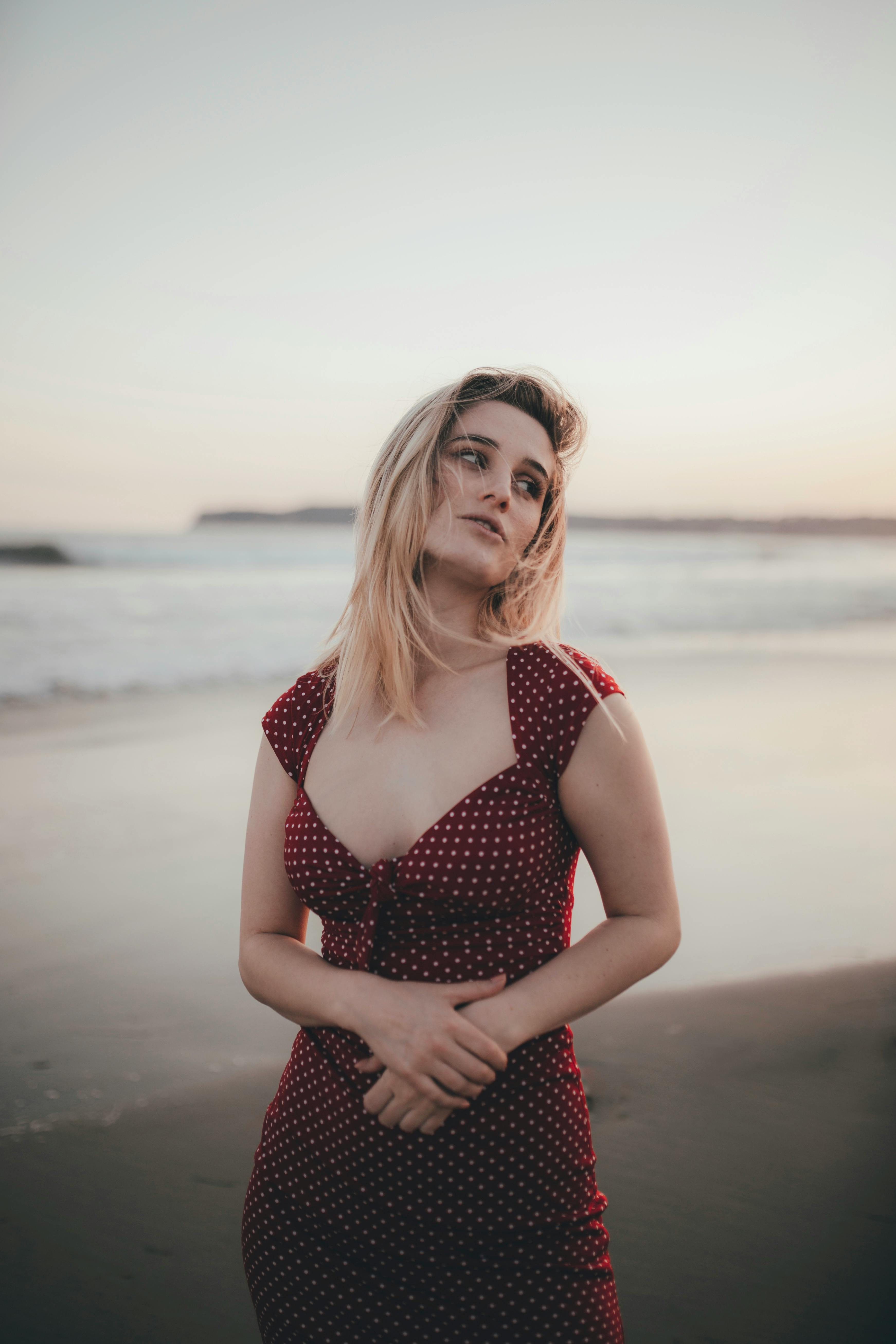A young woman poses in a red polka dot dress on a serene beach during sunset.