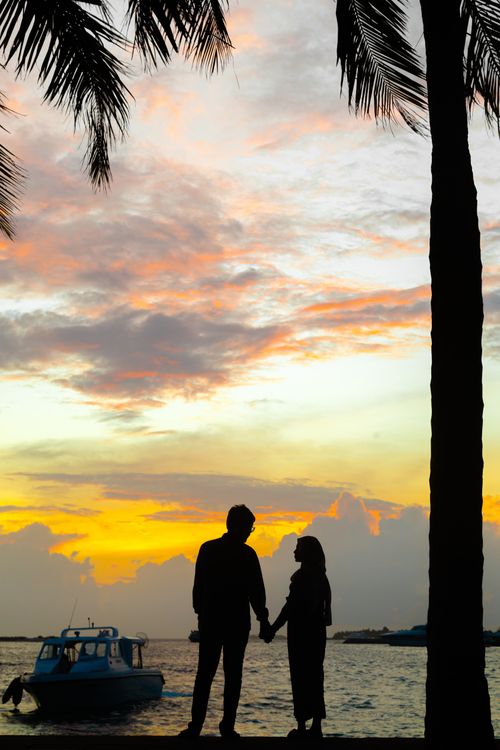 Free Silhouette of couple holding hands by the sea during sunset with palms and boat in view. Stock Photo