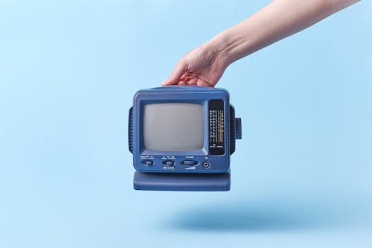 A hand holding a vintage blue CRT television against a light blue background. Studio shot.