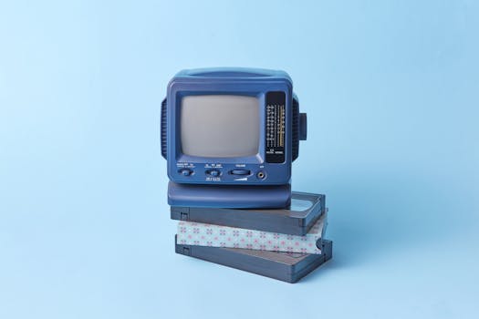 Vintage blue TV set featured atop VHS tapes on a pastel blue background, evoking a retro vibe.