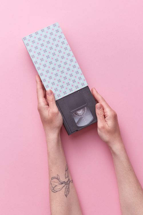 Free A VHS tape with floral cover held by tattooed hands on a pastel pink background, evoking nostalgia. Stock Photo