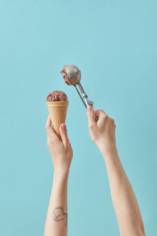 Free A hand holding a chocolate ice cream cone with a metal scoop above. Minimalist food photography. Stock Photo