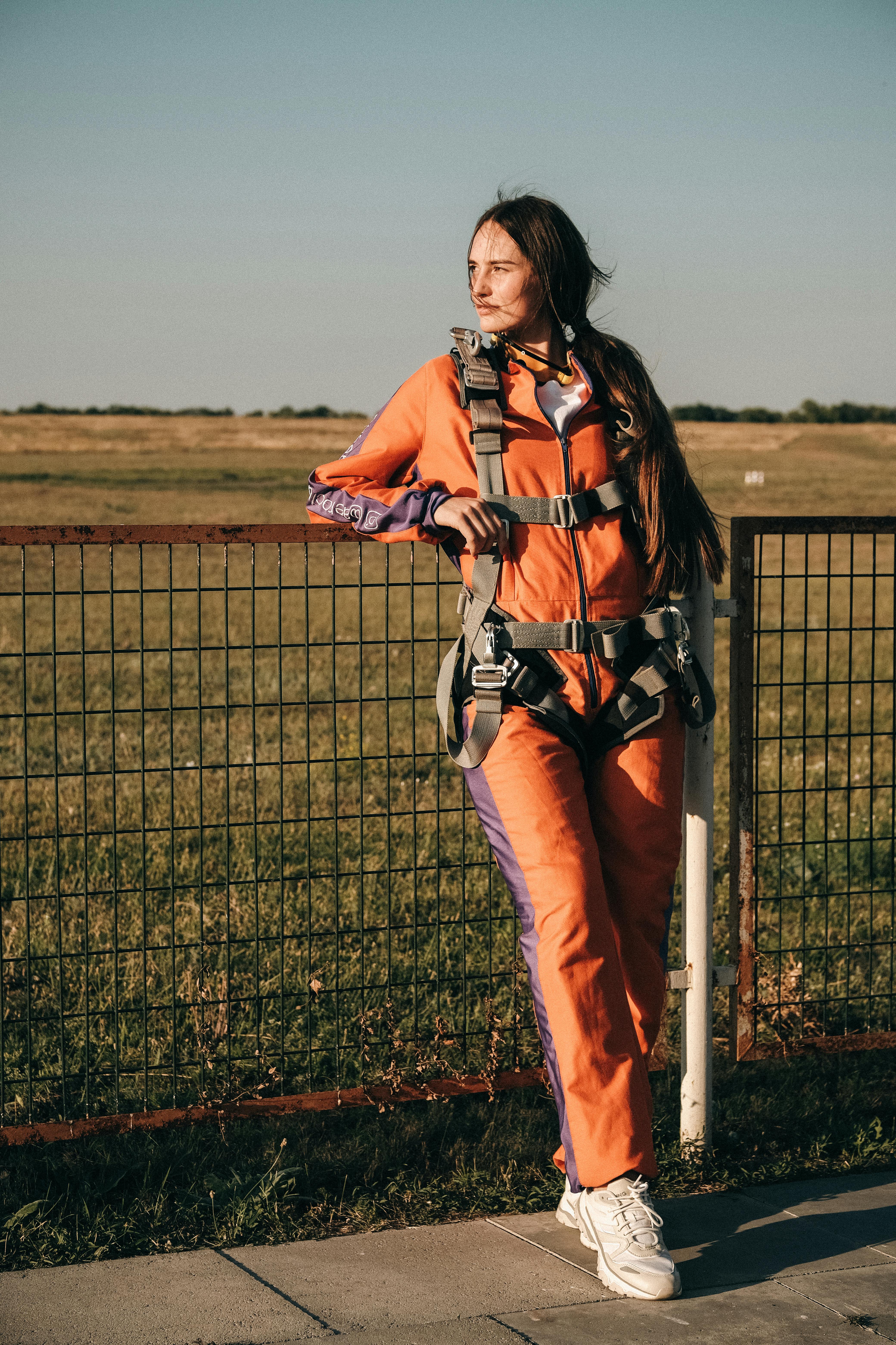 Young dreamy female in skydiving jumpsuit standing on pavement while leaning on grid fence and looking away against airdrome