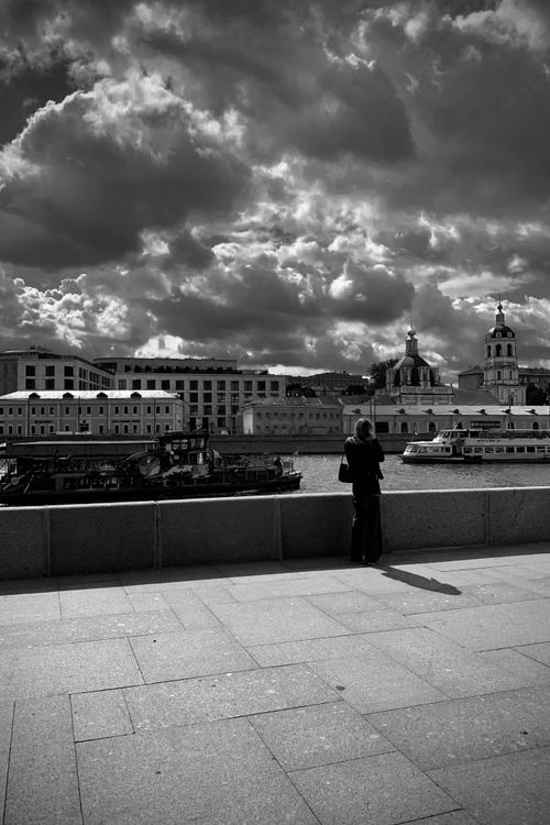 Free Black and white photo of a woman observing a riverfront with boats and historic buildings under dramatic clouds. Stock Photo