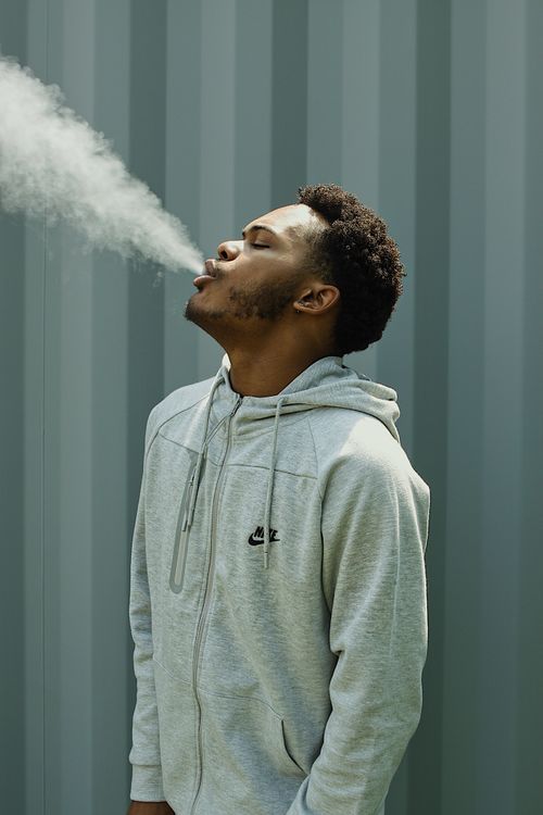 Free Young African American man in a hoodie exhaling smoke outdoors, highlighting relaxation and urban style. Stock Photo