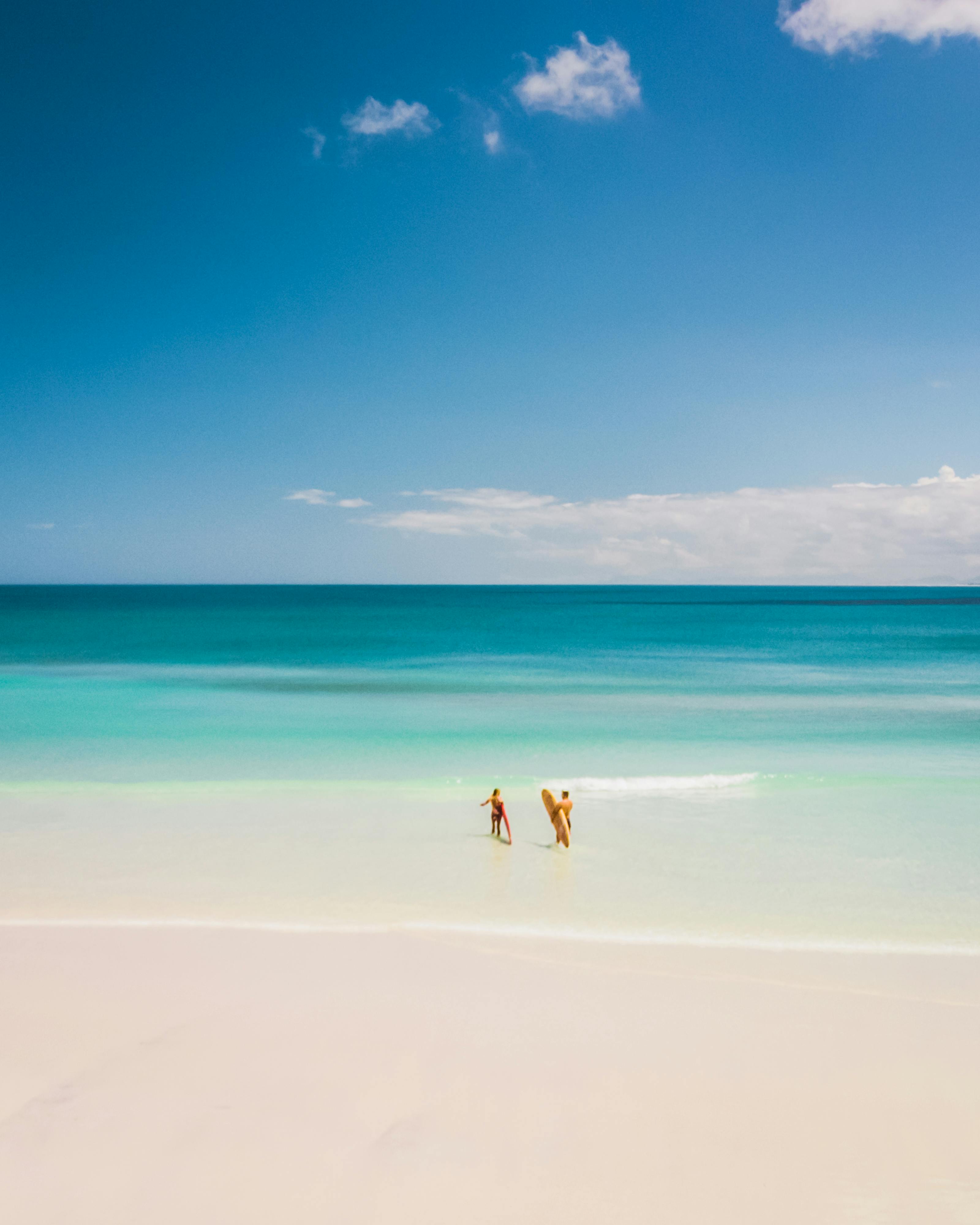 A serene beach scene with two people walking along clear blue waters under a bright sky.