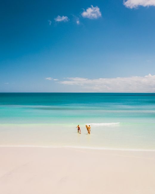 A serene beach scene with two people walking along clear blue waters under a bright sky.