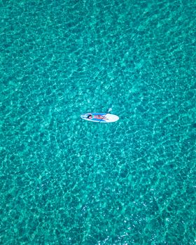 High-angle view of a woman paddleboarding in a vast, clear turquoise sea on a sunny day.
