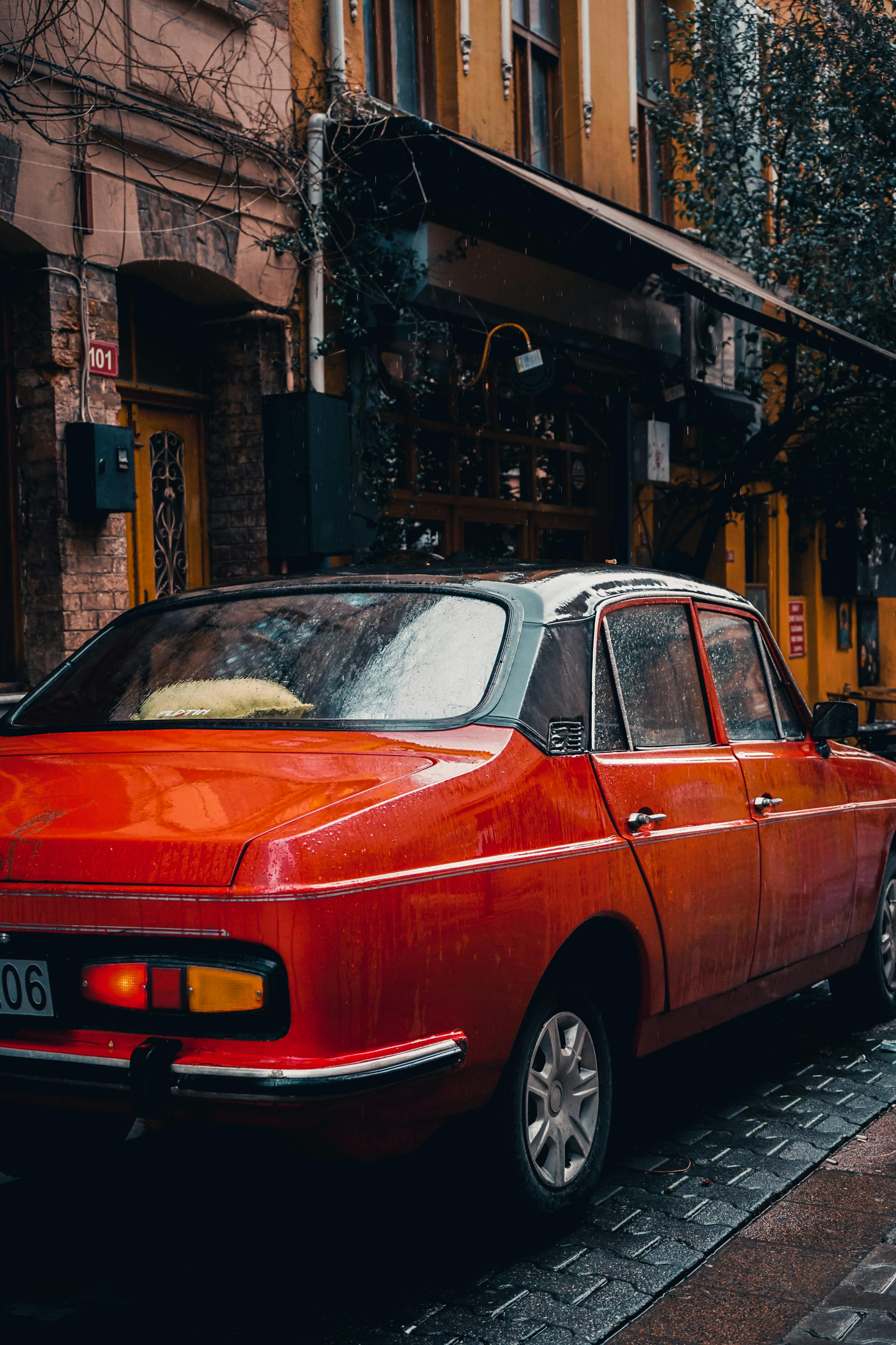 Free Classic red sedan parked on a wet cobblestone street, evoking a vintage urban feel. Stock Photo