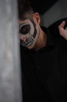 Moody portrait of a man with skull face paint leaning against a wall, capturing a spooky Halloween vibe.