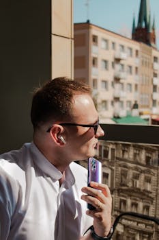 Man using wireless earbuds and smartphone in a sunny cityscape with modern architecture.
