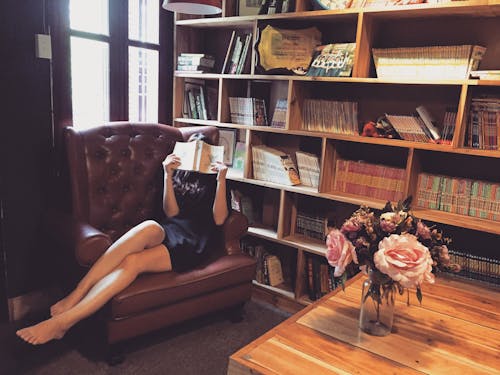 黑色迷你连衣裙的女人坐在棕色木制书架旁边的棕色皮革簇绒的沙发椅子上