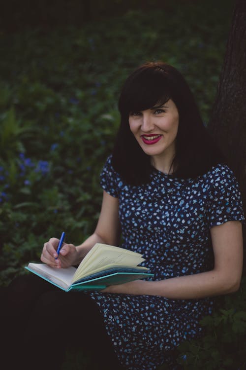 Mulher Em Camisa De Manga Curta Floral Branca E Azul Com Gola Redonda Segurando Um Caderno Azul Petróleo Apoiado Em Troncos Marrons