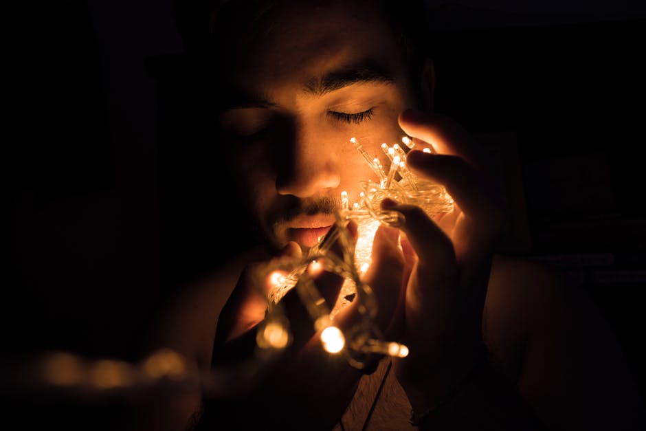 A man closes his eyes while holding glowing string lights in a dark setting.