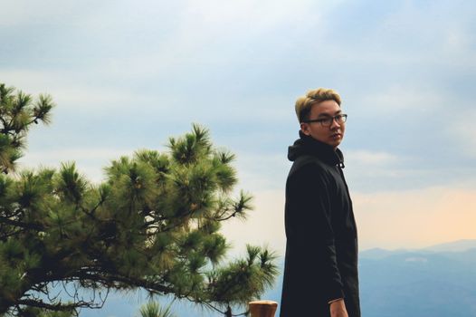 A young man in glasses stands outdoors near a pine tree with mountains in the background.