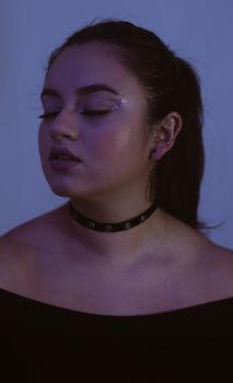 A serene portrait capturing a woman with closed eyes, wearing glitter makeup and a choker, in soft lighting.