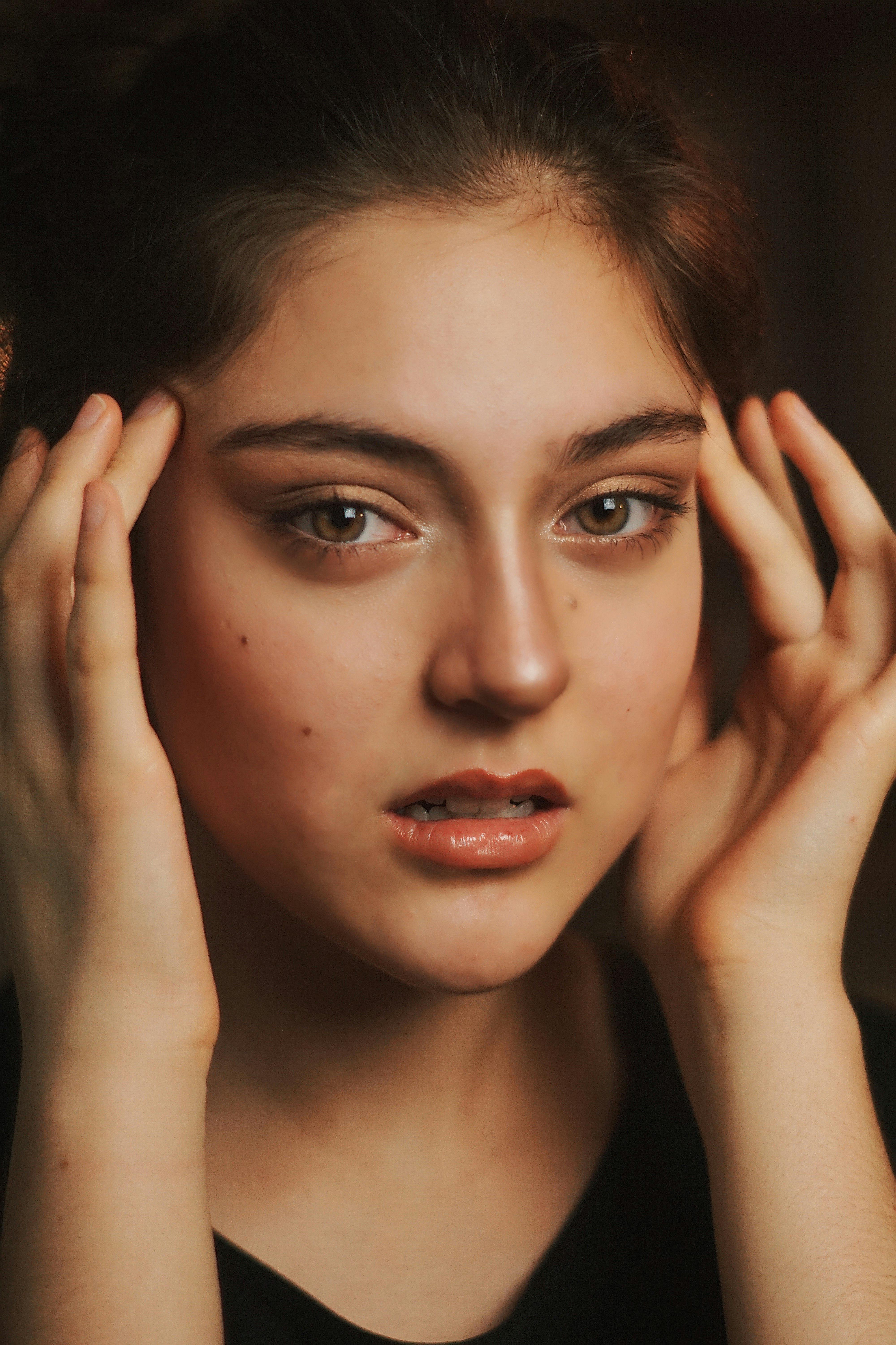 Close-up portrait of a woman with brown eyes, hands on face, looking contemplative.