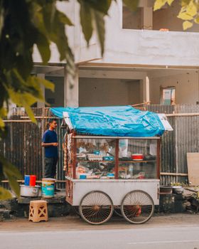 Street vendor standing beside a food cart in an urban area with a building behind.