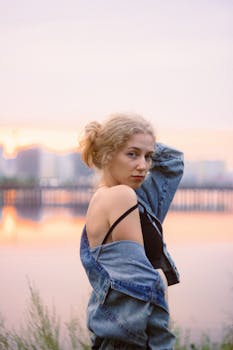 A blonde woman in a denim jacket poses by the waterfront during a colorful sunset.