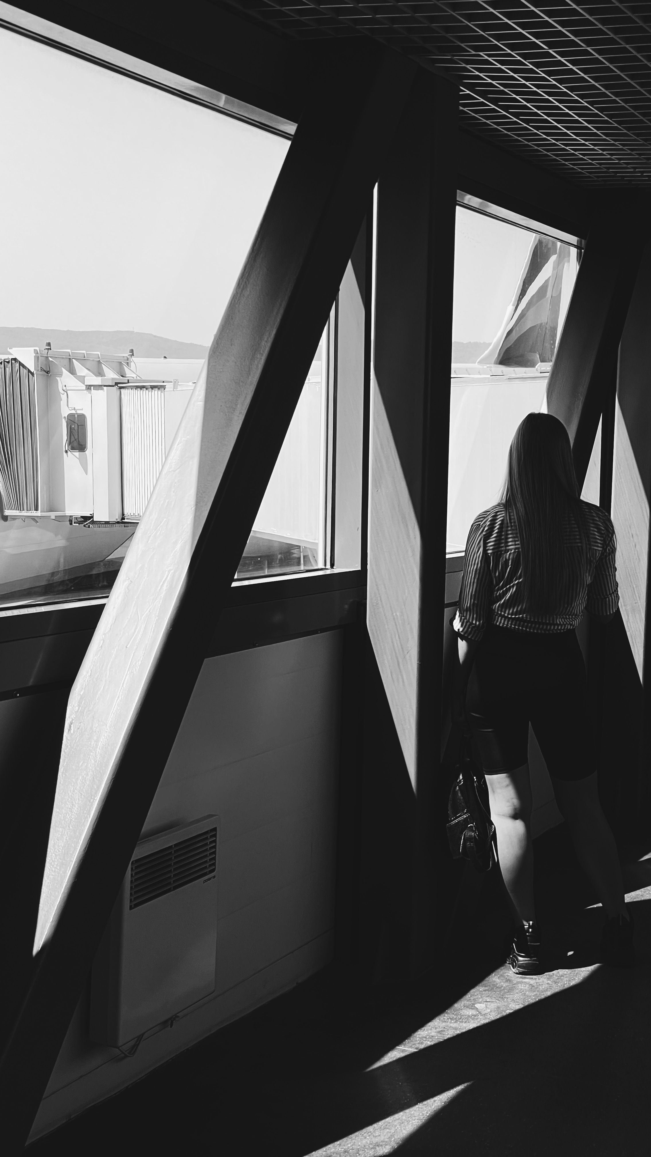 Black and white photo of a woman viewing an airport runway through large windows.