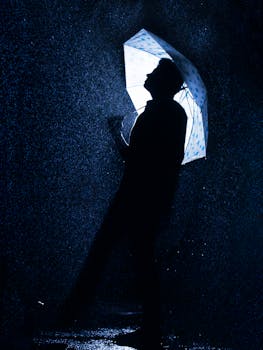 A silhouette of a man holding an umbrella under rainfall at night, creating a dramatic effect.