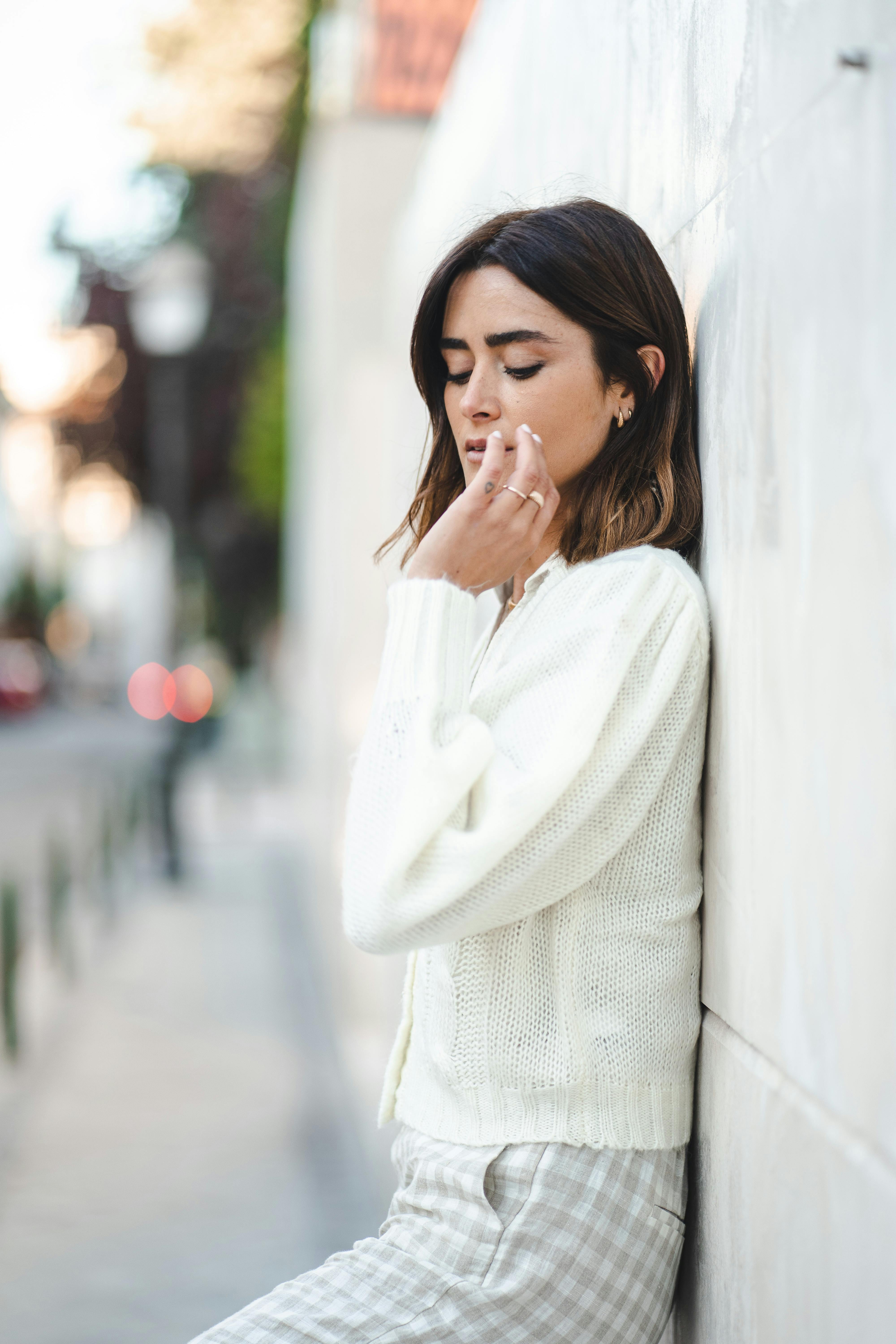 Contemplative woman in a white cardigan leans against a wall on a city street during daytime.