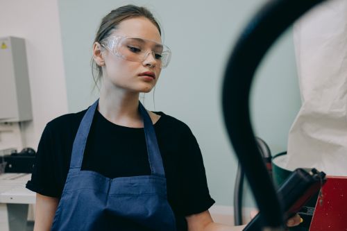 Free Young woman in a workshop setting, wearing protective goggles and a blue apron, standing indoors. Stock Photo