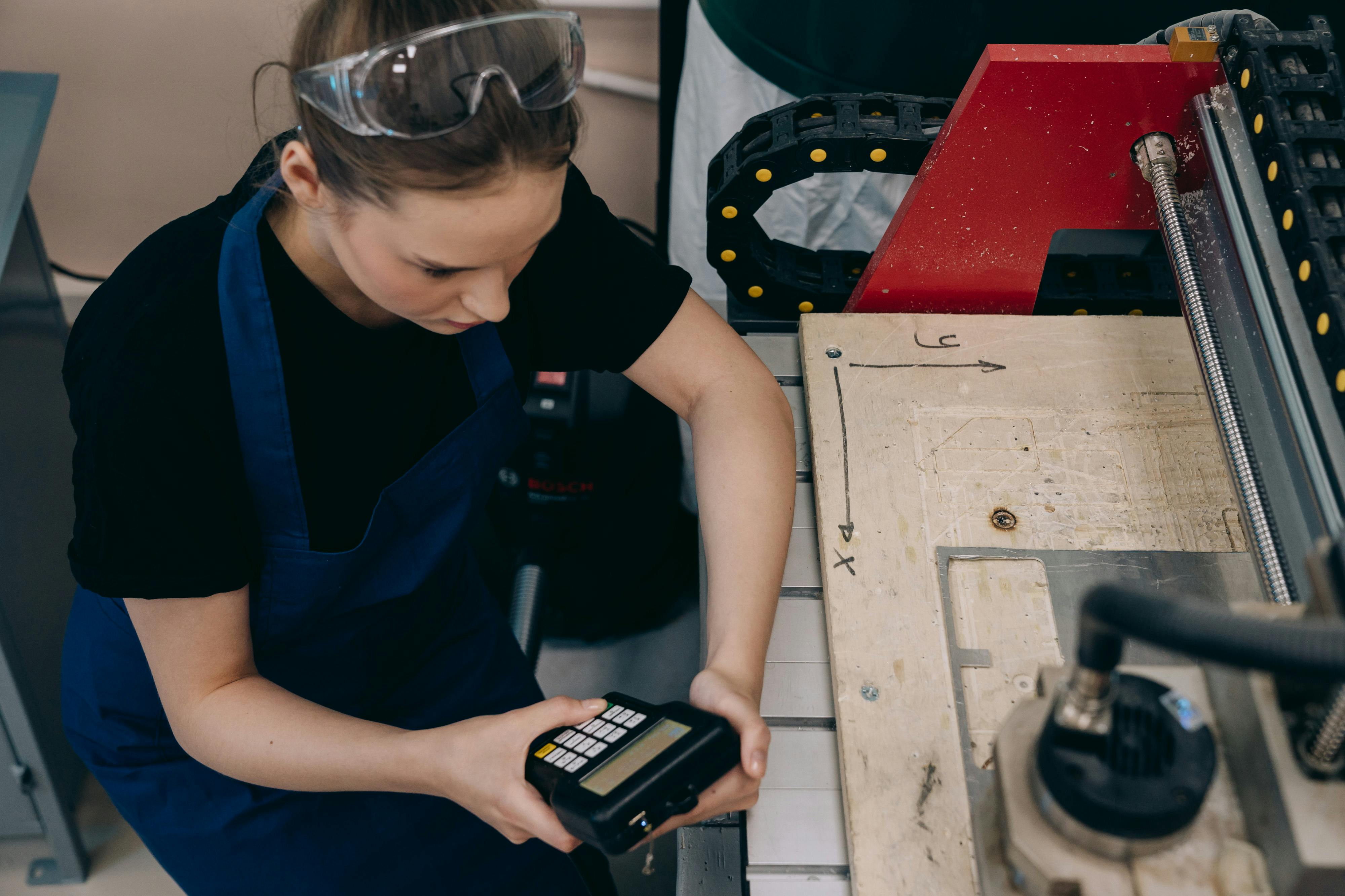 Woman in workshop using control device on machinery, wearing protective goggles and apron.