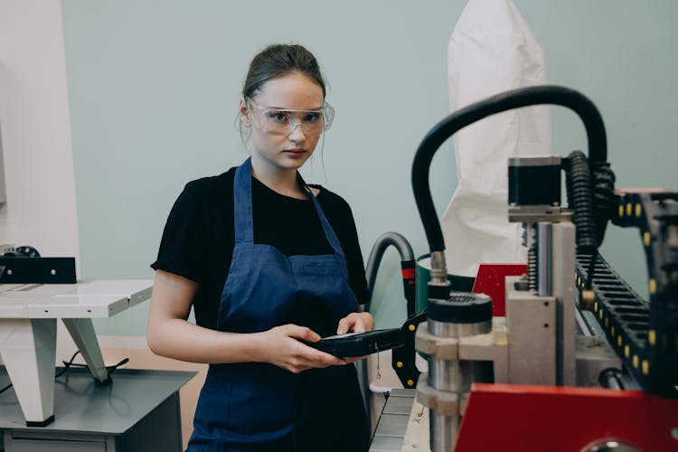 A Young Woman In An Apron And Safety Glasses Standing By A CNC Machine