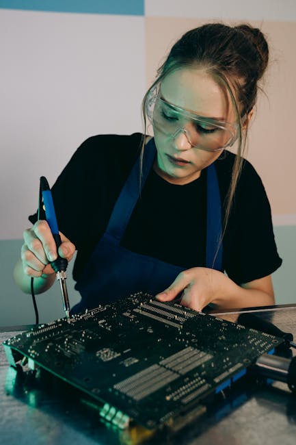 A young woman in an apron and goggles soldering a circuit board indoors.