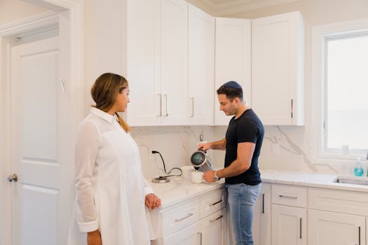 A couple standing in a modern kitchen, one pouring a drink, in a bright and cozy setting.