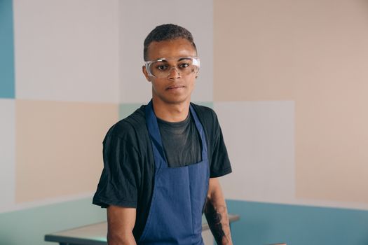 Young man wearing an apron and safety glasses in a workshop setting.