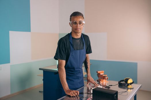 Young technician in apron and safety glasses standing at a workstation indoors, ready for work.
